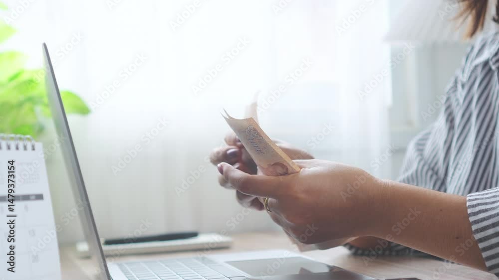 Close-up of a woman's hands counting Thai baht money in front of a laptop, showing the concept of earning money online, working from home, or making digital income