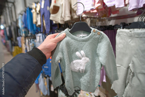 Hand holding soft green baby sweater with cute bunny design in cozy retail store while browsing through display of knit clothing for infants. Retail display showcasing variety of baby sweaters