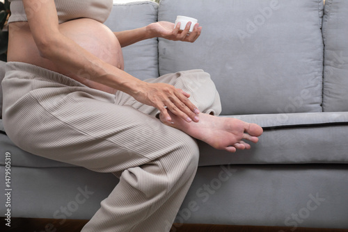 Asian pregnant woman sitting on sofa applying lotion to her foot with gentle care, promoting relaxation and skin comfort during pregnancy in a cozy home setting.