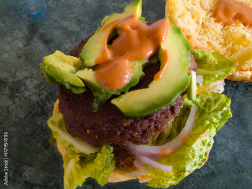 Burger with beef patty on lettuce topped with avocado slice and burger sauce served on green marble chopping board 