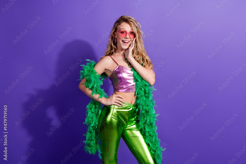 Naklejka premium Trendy young woman in vibrant outfit posing on a violet background, showcasing retro style with glowing top and green boa.