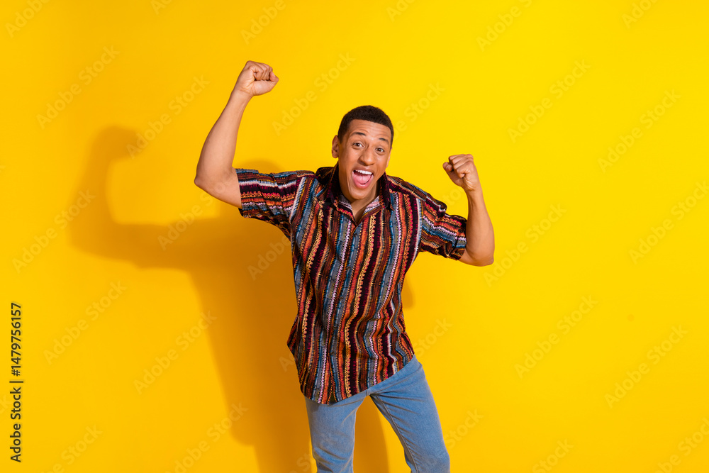 Naklejka premium Young man celebrating with joy against a vibrant yellow background while wearing a colorful casual shirt and denim jeans