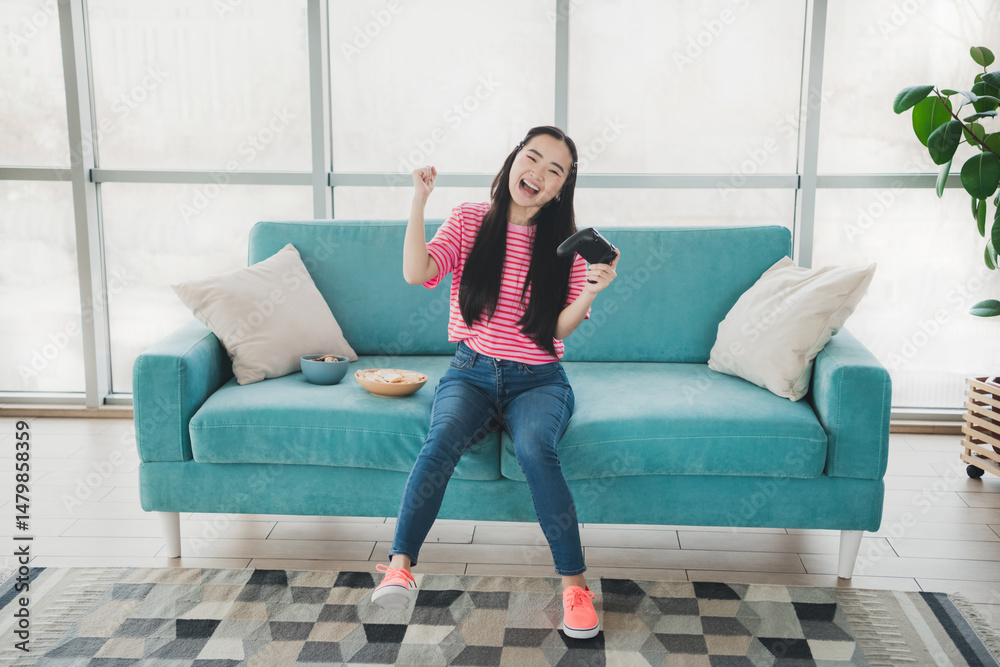 Obraz premium Young woman smiling while gaming on a turquoise couch in a modern living room with snacks and natural daylight