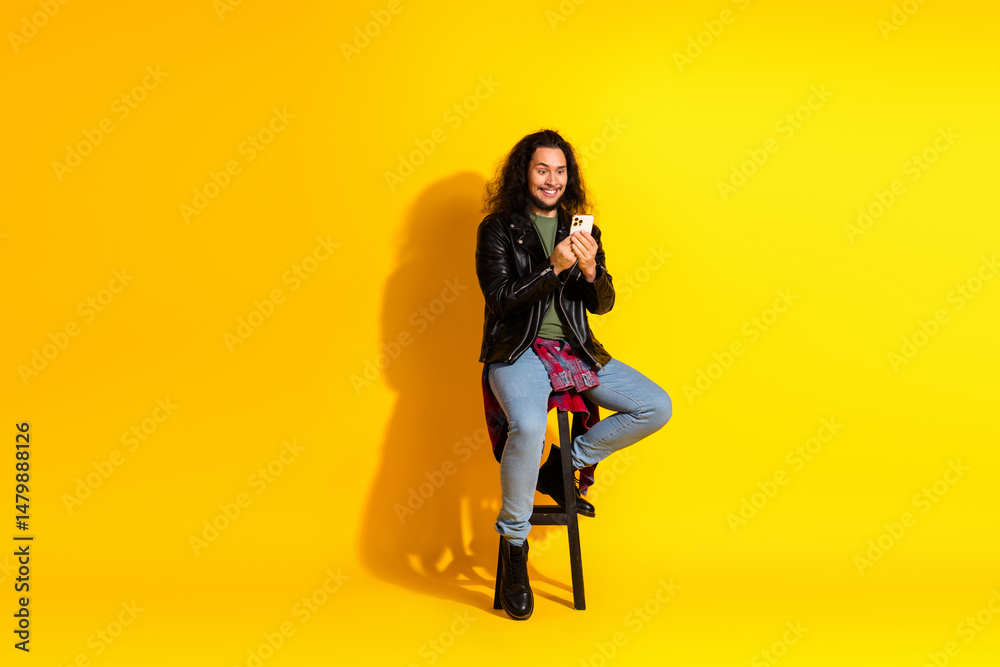 Obraz premium Young stylish man in a leather jacket sitting on stool holding a smartphone against yellow background.