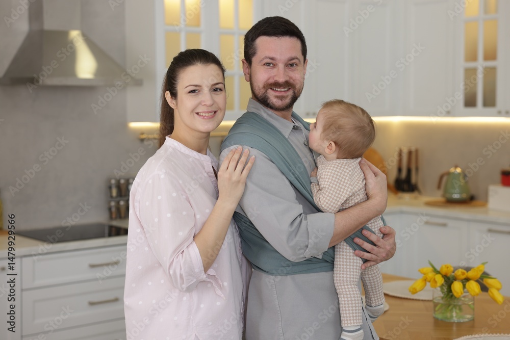 Fototapeta premium Portrait of happy family indoors. Father holding his child in sling (baby carrier) at home