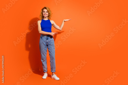 Cheerful young woman with red hair wearing a stylish outfit smiling and gesturing against a bright orange background expressing positivity