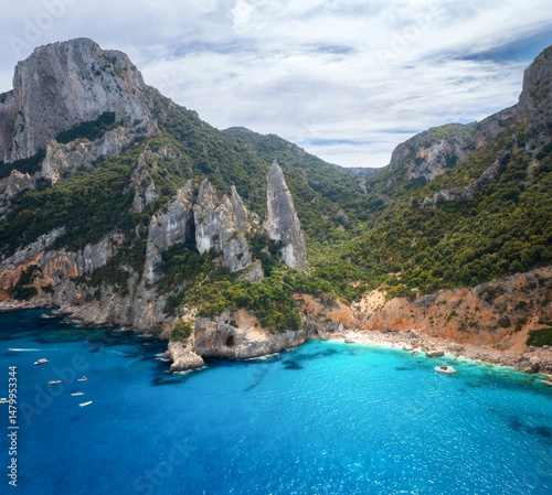 Aerial view of rocks and mountains, blue bay, beach, boats on sunny summer day. Cala Goloritze, Sardinia Island, Italy. Top drone view of cliffs, stones, green trees, transparent azure water at sunset