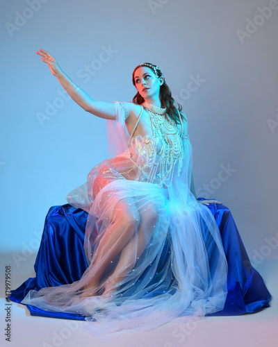 Fantasy studio portrait of a red-haired mermaid faery model wearing a white pearl gown, fairytale costume. posing in a classical sitting position with flowing fabric and ethereal studio lighting