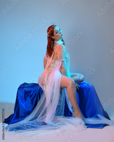 Fantasy studio portrait of a red-haired mermaid faery model wearing a white pearl gown, fairytale costume. posing in a classical sitting position with flowing fabric and ethereal studio lighting