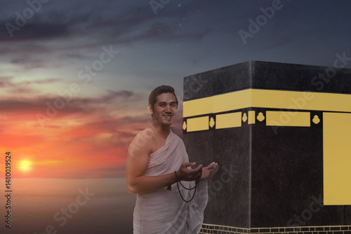 Indonesian southeast asian muslim man wearing white traditional clothes or ihram raises his hand and prays while holding prayer beads in front of the Kaaba. Concept of hajj or umrah