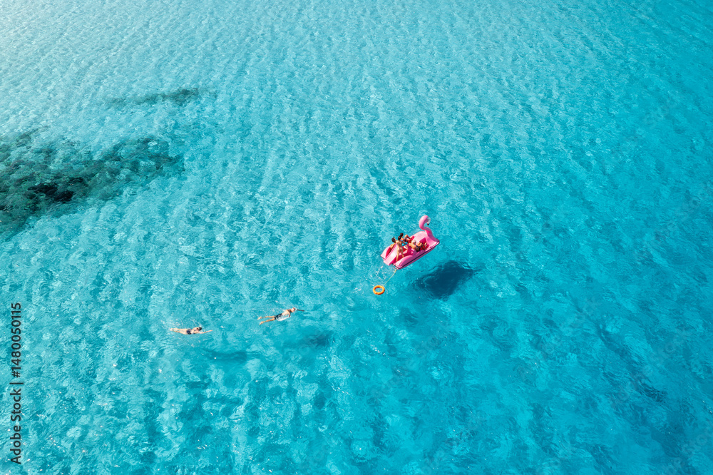 Obraz premium Aerial view of Inflatable pink flamingo with people on crystal clear blue water on summer sunny day. Top drone view of relax family on boat, azure sea in Sardinia Island, Italy. Travel and vacation