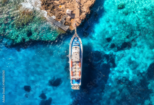 Aerial view of boat with people and rocky sea coast on sunny summer day. Top drone view of yacht, transparent azure water, path. Mediterranean sea. Travel in Sardinia island, Italy. Tropical landscape