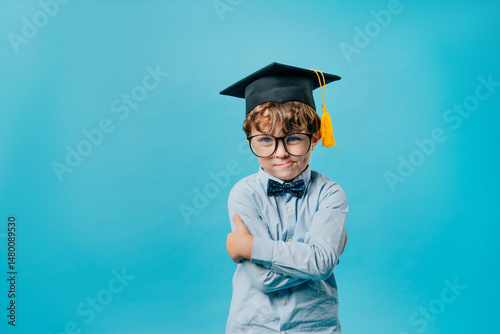 Emotional preschool boy wearing graduation cap, glasses and bow tie over blue background