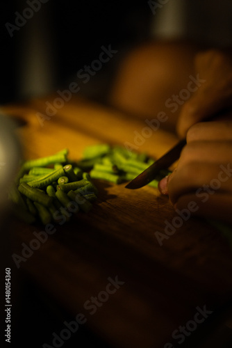 Hands slice fresh green beans on a wooden board, capturing a quiet, warm moment of simple, focused home cooking.