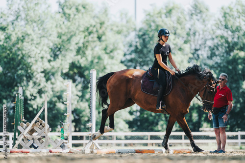 A rider on a horse practices jumping outdoors with guidance from a coach. The scene depicts focus and teamwork in an equestrian training setting amidst lush greenery.