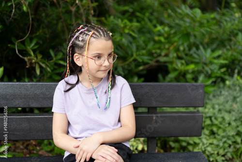 Little girl with glasses and dreadlocks smiling sits on a bench in a summer park. Concept of children's leisure and fun