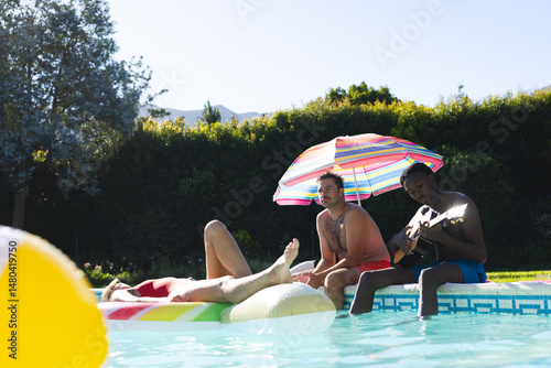 Relaxing Diverse friends in swimwear soaking up sun at backyard pool, with rainbow float and guitar