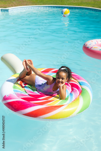 Relaxing African American girl floating on rainbow tube in backyard pool, with doughnut float