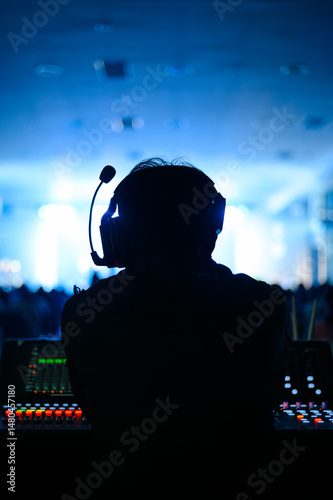 Silhouette of a sound technician wearing a headset, working at a control panel during a live event. The background features blue stage lighting, creating a vibrant atmosphere.	