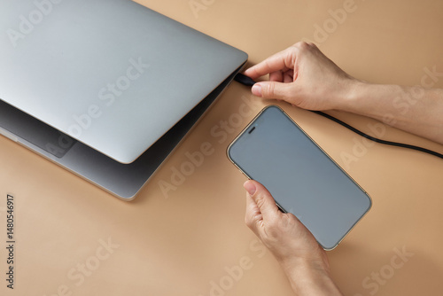 female hand connecting charger cable to laptop and holding smartphone at beige table, closeup