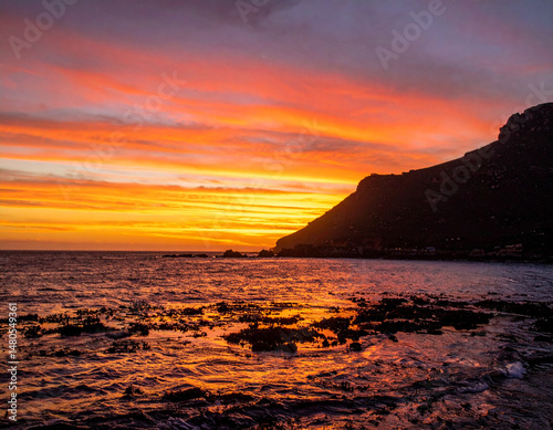 A South African sunset over a coastal cliff with deep orange skies reflecting on water. photo