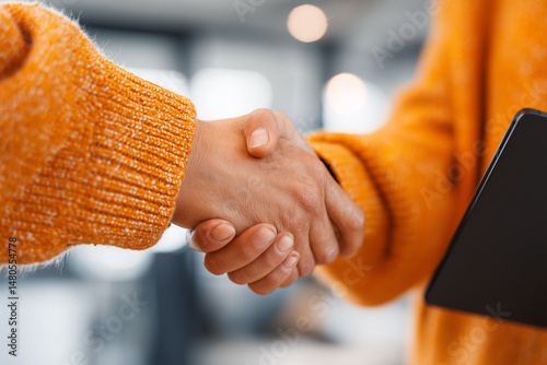 Close-up of two business people shaking hands and closing a deal, wearing orange sweaters and holding black digital tablets in an office