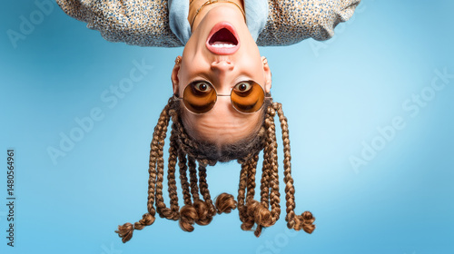Young woman wearing sunglasses and braided hair hanging upside down with a surprised expression on blue background
