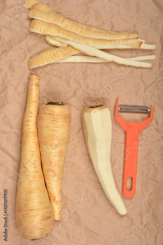 Peeled and unpeeled parsnips lie on a brown paper surface next to an orange vegetable peeler