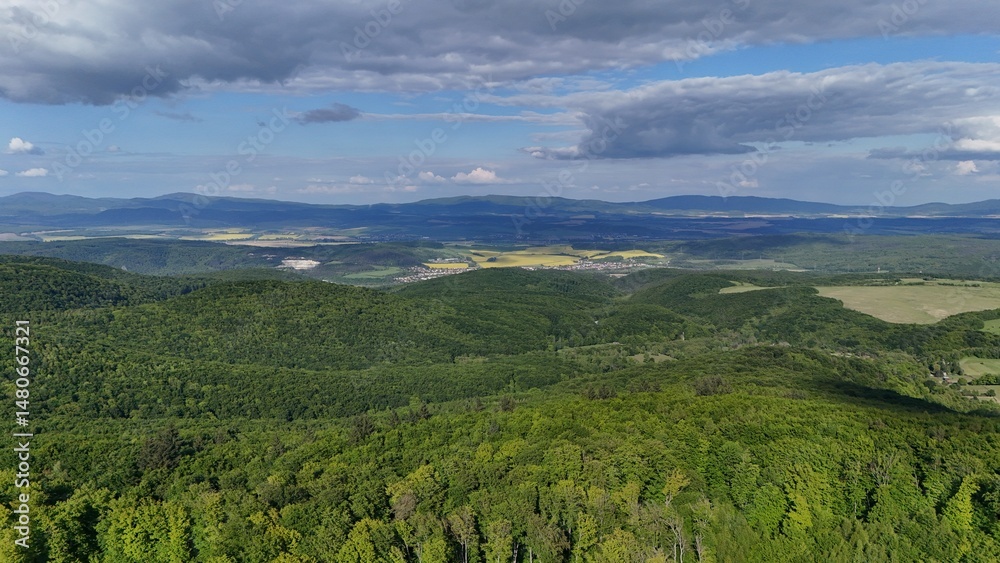 Obraz premium Aerial view of scenic forest-covered mountains under a sunny sky with soft clouds. Stunning natural landscape captured by drone, perfect for travel, nature, and environment themes.