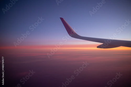 An airplane wing is captured against a stunning twilight sky filled with a gradient of blue purple orange and pink, symbolizing air travel and the serene beauty of a sunset from above the clouds.