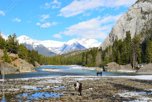 Banff, AB, Canada - April 14, 2025 : Tourists resting on the riverside promenade of the Bow Falls Trail in the Banff National Park, Alberta, Canada. This was on a cold sunny spring afternoon. 		