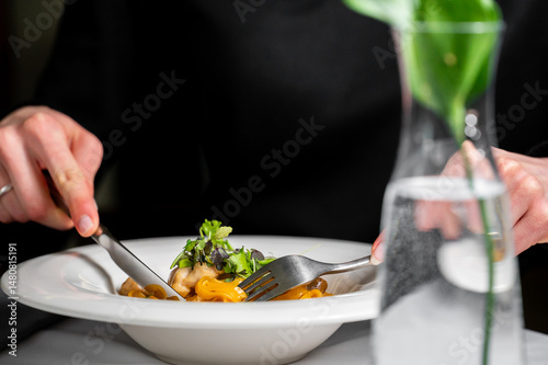 A person enjoys a plate of pasta with fresh greens, using a fork and knife. The elegant setting includes a vase with water and a green plant, creating a refined dining atmosphere.