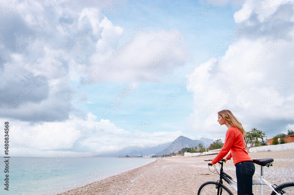 Fototapeta premium Young Woman with Bicycle by the Beach on a Cloudy Day..