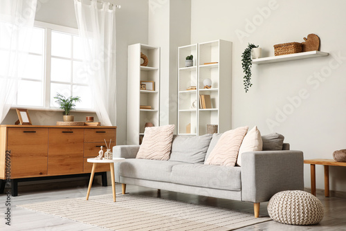 Shelving unit, pouf and grey sofa near white wall in interior of living room