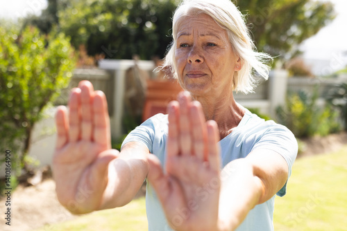 Senior female performing forward arm stretch in sunny backyard garden, practicing gentle exercise photo