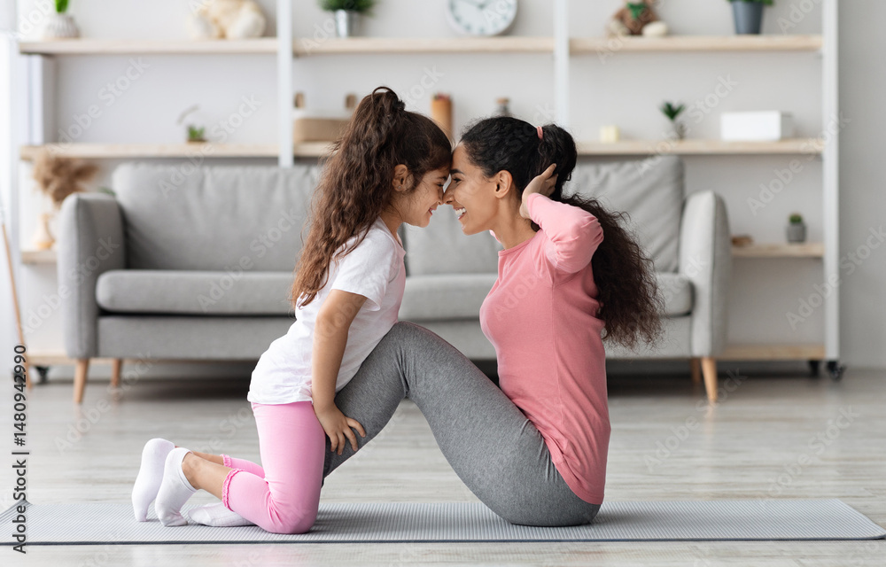 Obraz premium Loving middle eastern mother and daughter in sportswear working on abs together, exciting little girl holding her mom legs while she doing crunches, touching each other with foreheads, home interior