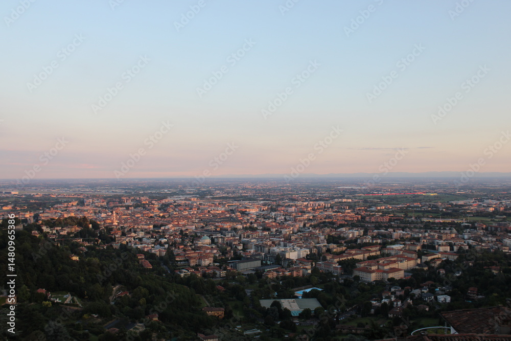 Panoramic view from Città Alta in Bergamo: the city stretches out in the warm sunset light, with rooftops, historic buildings, and flatlands fading into the horizon under a clear sky. Panoramic view from Città Alta in Bergamo: the city stretches out in the warm sunset light, with rooftops, historic buildings, and flatlands fading into the horizon under a clear sky.