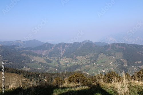 Panorama from the summit of Monte Brozzone: green valleys dotted with houses, forested mountains, and distant peaks fading into the haze under a clear and serene sky.