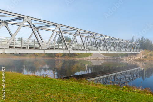 A pedestrian deck with metal railings spans a tranquil, narrow river. The bridge adjoins two riverbanks. Steam rises from the water, reflecting the structure in the water. 