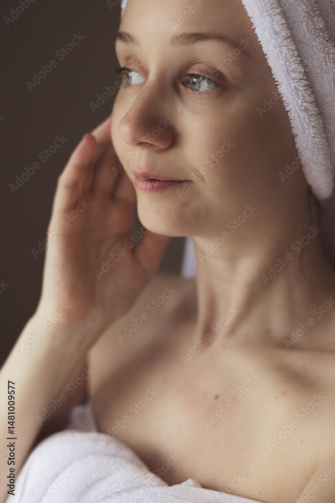 Fototapeta premium A woman with a towel on her head, after a bath, looks away with a gentle expression. A woman wrapped in a white towel after a bath. Spa day relaxing.