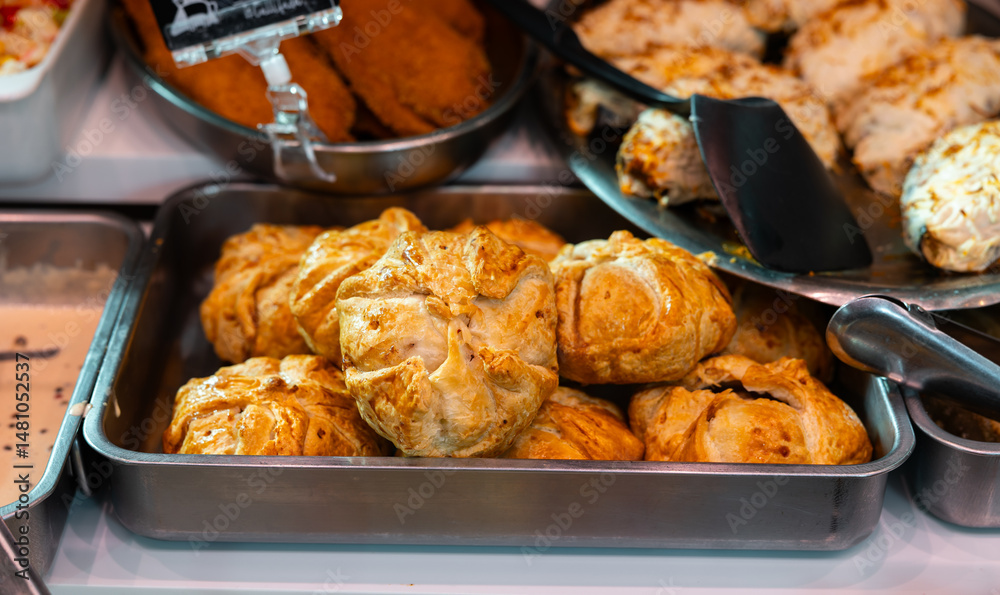 Traditional Spanish golden baked empanadas, with crispy olive oil crust, filled with paprika seasoned beef, sweet red pepper, and boiled eggs, displayed on metal tray in deli section of local store