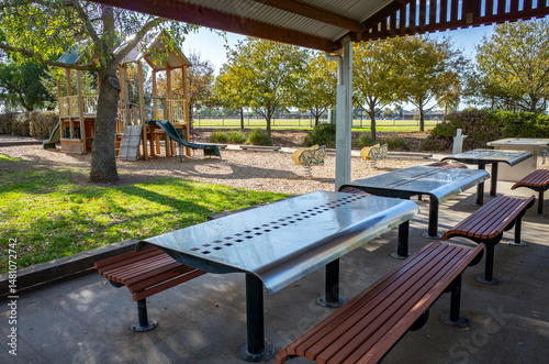Covered picnic shelter with barbecue facilities, dining tables,and seating benches in an Australian public park, next to a children's playground—family recreation, social gathering and outdoor leisure