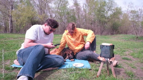 couple and their dog on picnic , cooking marshmallows on camping stove and using smartphone
