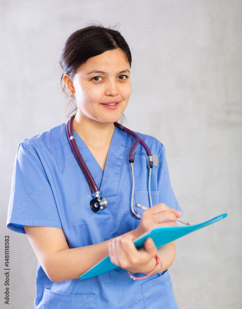 Positive polite young woman general practitioner in blue uniform and with phonendoscope around neck filling out clipboard with medical records. Studio photoshoot on gray background