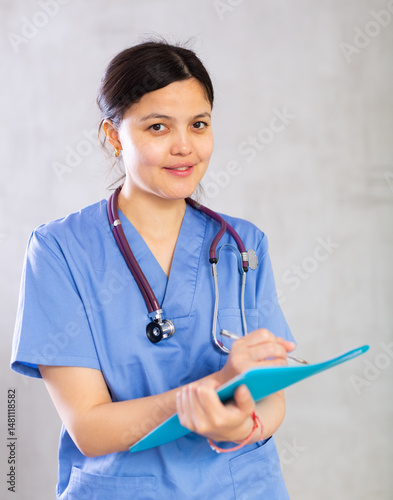 Obraz na płótnie Positive polite young woman general practitioner in blue uniform and with phonendoscope around neck filling out clipboard with medical records. Studio photoshoot on gray background w salonie