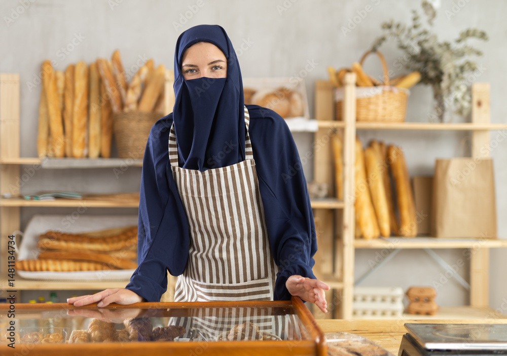 European woman in hijab offering fresh pastries behind the counter. Muslim bakery owner stands in front of bread.