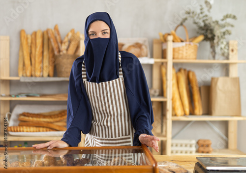 Obraz na płótnie European woman in hijab offering fresh pastries behind the counter. Muslim bakery owner stands in front of bread. w salonie