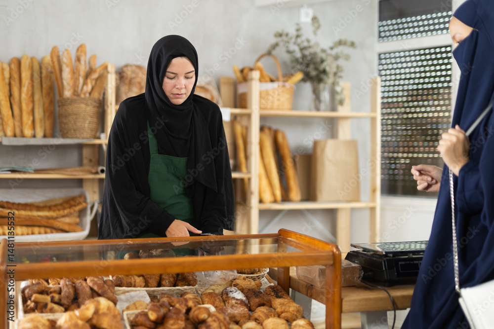 Focused young Muslim woman of European appearance wearing black hijab working in cozy small bakery, arranging freshly baked pastries on display