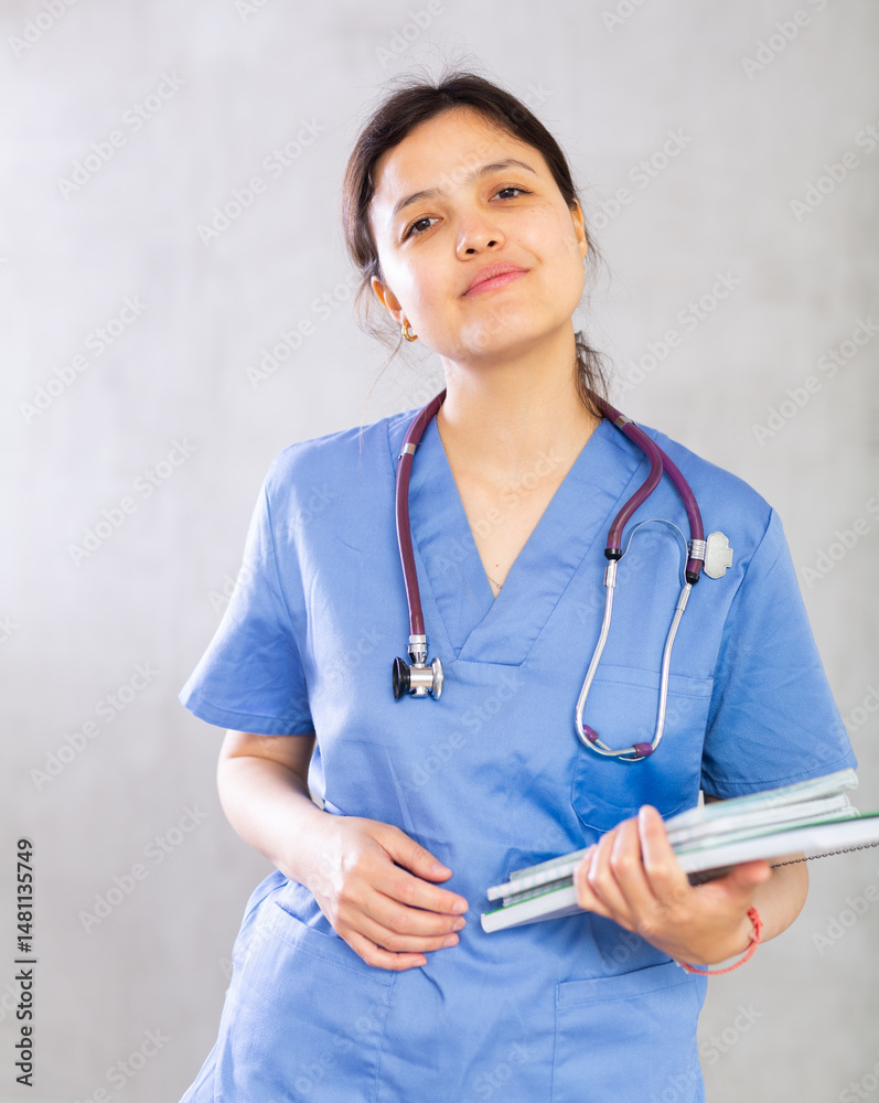 Positive confident young woman, professional doctor, in blue uniform and with phonendoscope around neck holding medical records. Studio photoshoot on gray background