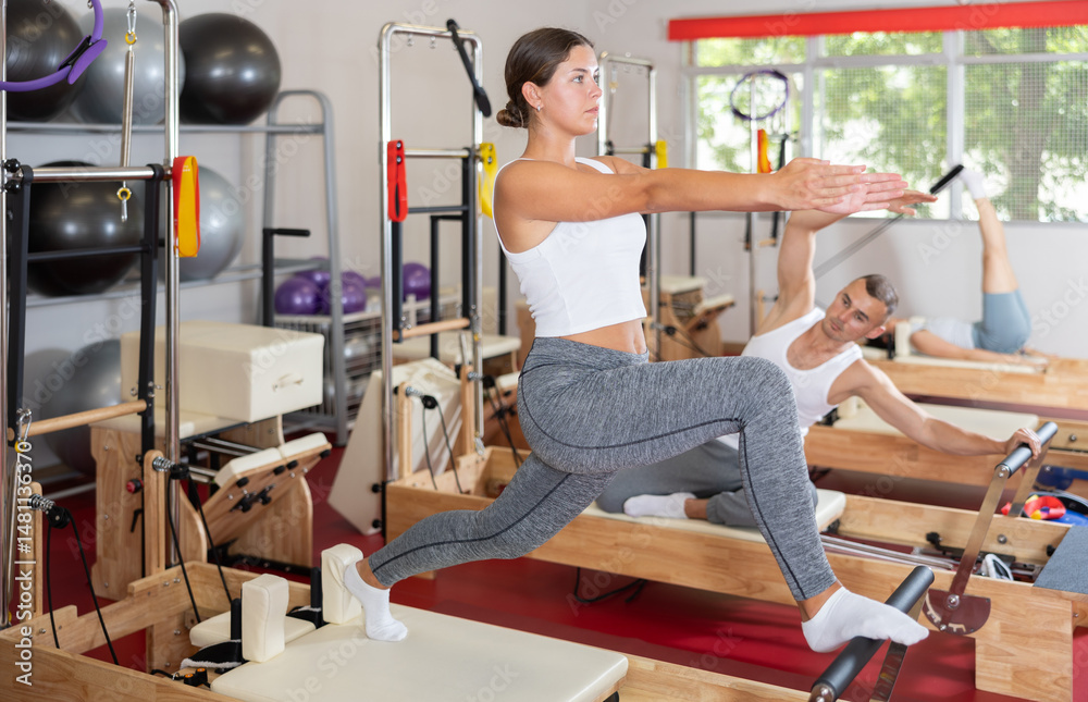 Sportive young girl training her arms and legs on reformer machine in fitness hall